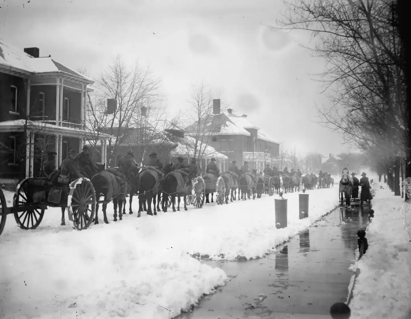 Horse-drawn wagons on a snow-covered street lined with houses, around 1915